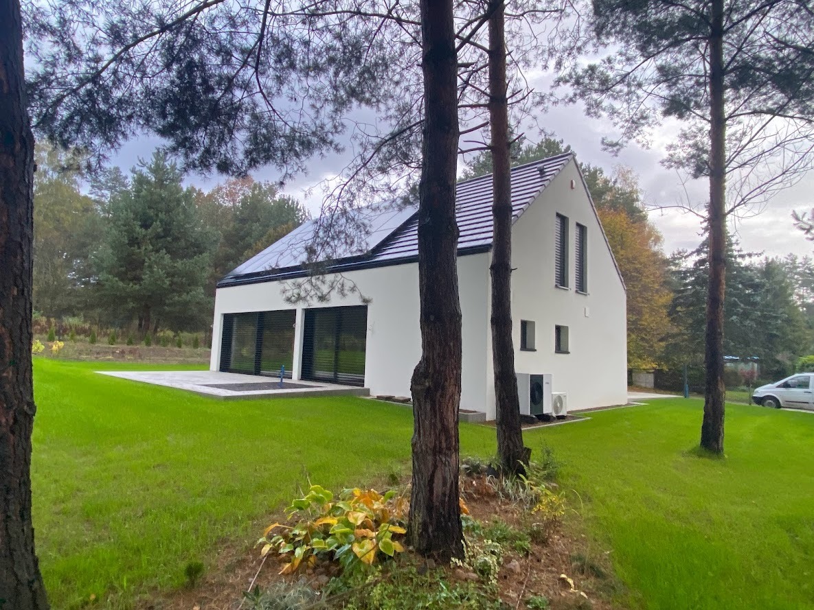 Nowoczesny, biały dom murowany z panelami słonecznymi na dachu, widoczny z perspektywy ogrodu, z drzewami na pierwszym planie i samochodem w tle.