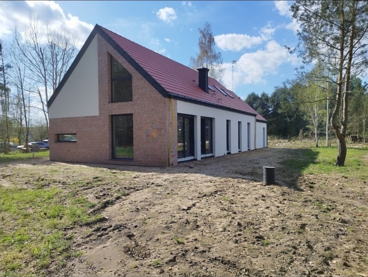 Nowoczesny dom jednorodzinny z czerwoną dachówką, elewacją z cegły i białym tynkiem, duże okna z czarnymi ramami, widok z zewnątrz na niezagospodarowany teren.