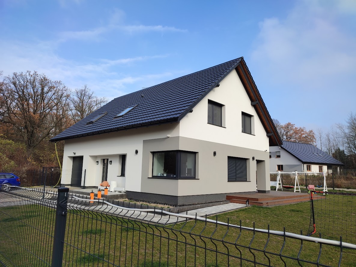 Nowoczesny dom jednorodzinny z ciemną dachówką i białymi ścianami, wykończony szarą elewacją w dolnej części, z widocznym tarasem i placem zabaw w ogrodzie oraz drugim budynkiem w tle.