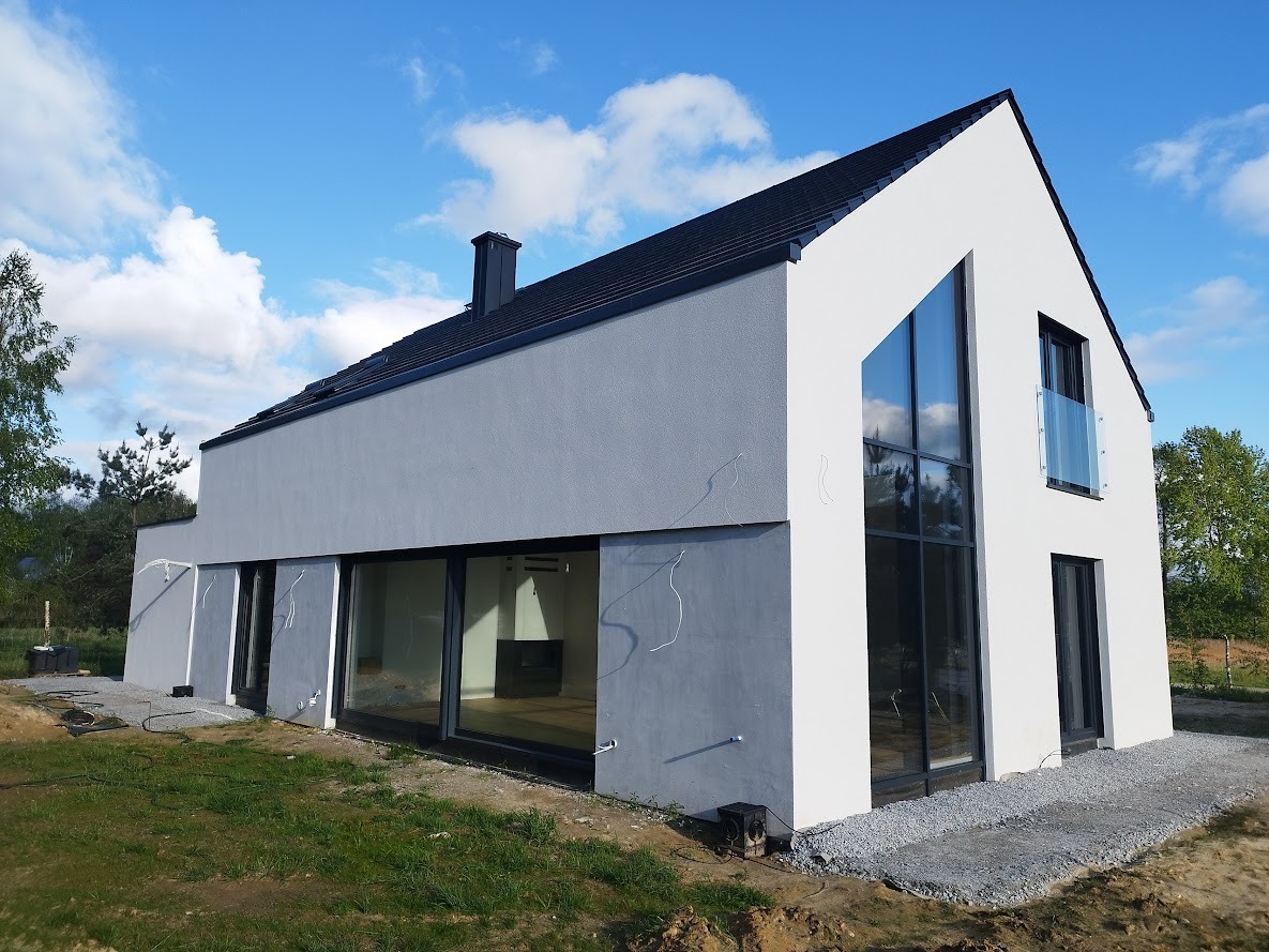 Nowoczesny dom jednorodzinny z dużymi przeszkleniami, wykończony w odcieniach szarości i bieli, z czarnym dachem i kominem. Widoczne okna panoramiczne i balkon z szklaną balustradą. Teren wokół...