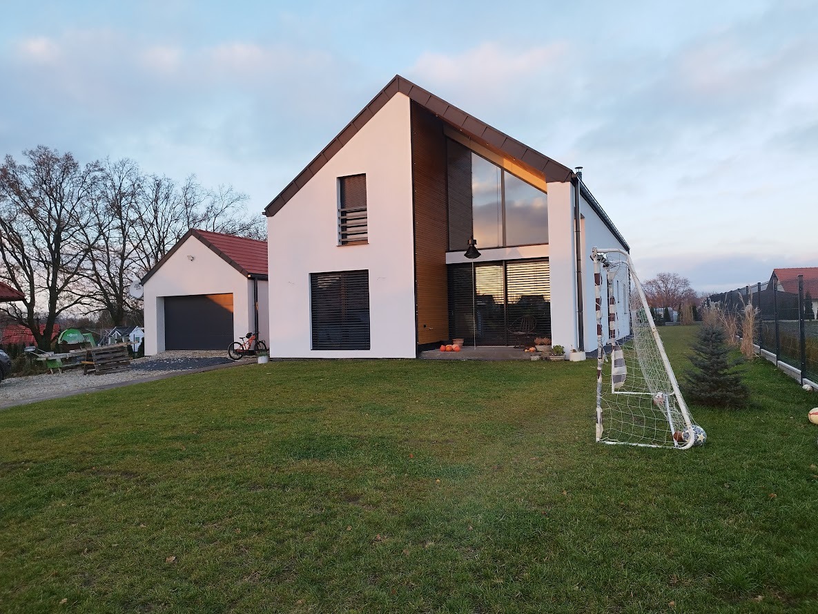 Nowoczesny dom jednorodzinny z dużymi oknami, garażem i bramą na pilota, oraz siatką do gry w piłkę na trawniku przed domem.