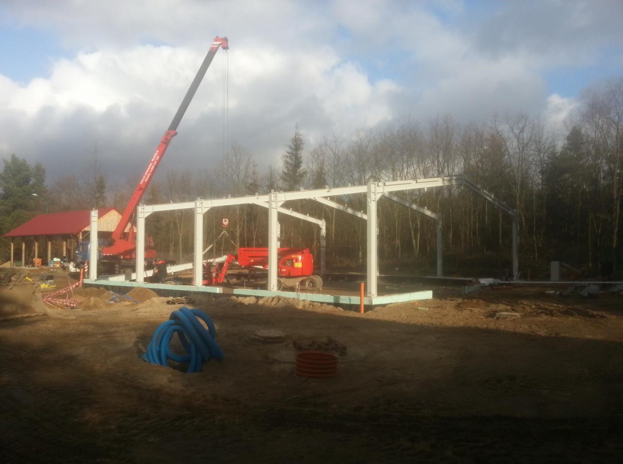 Montaż stalowej konstrukcji hali na fundamentach, widoczny żuraw, podnośnik koszowy i przygotowane rury instalacyjne.