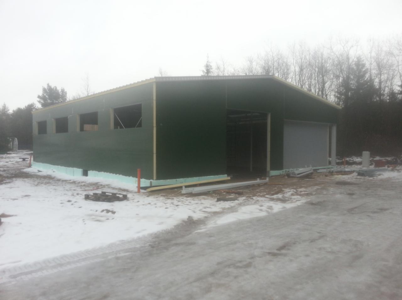 Budowa hali magazynowej z zielonej blachy w Obornikach Śląskich, widoczne ocieplenie fundamentów styropianem i częściowo zamontowana brama segmentowa.