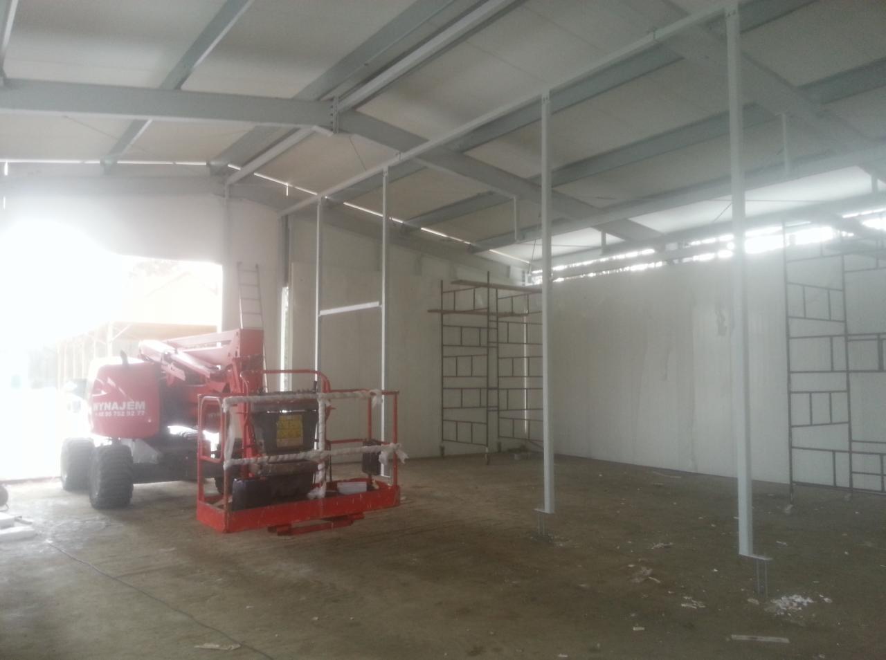 Wnętrze budowanego obiektu chłodniczego z widoczną konstrukcją stalową, ścianami z paneli warstwowych i podnośnikiem koszowym wewnątrz hali.