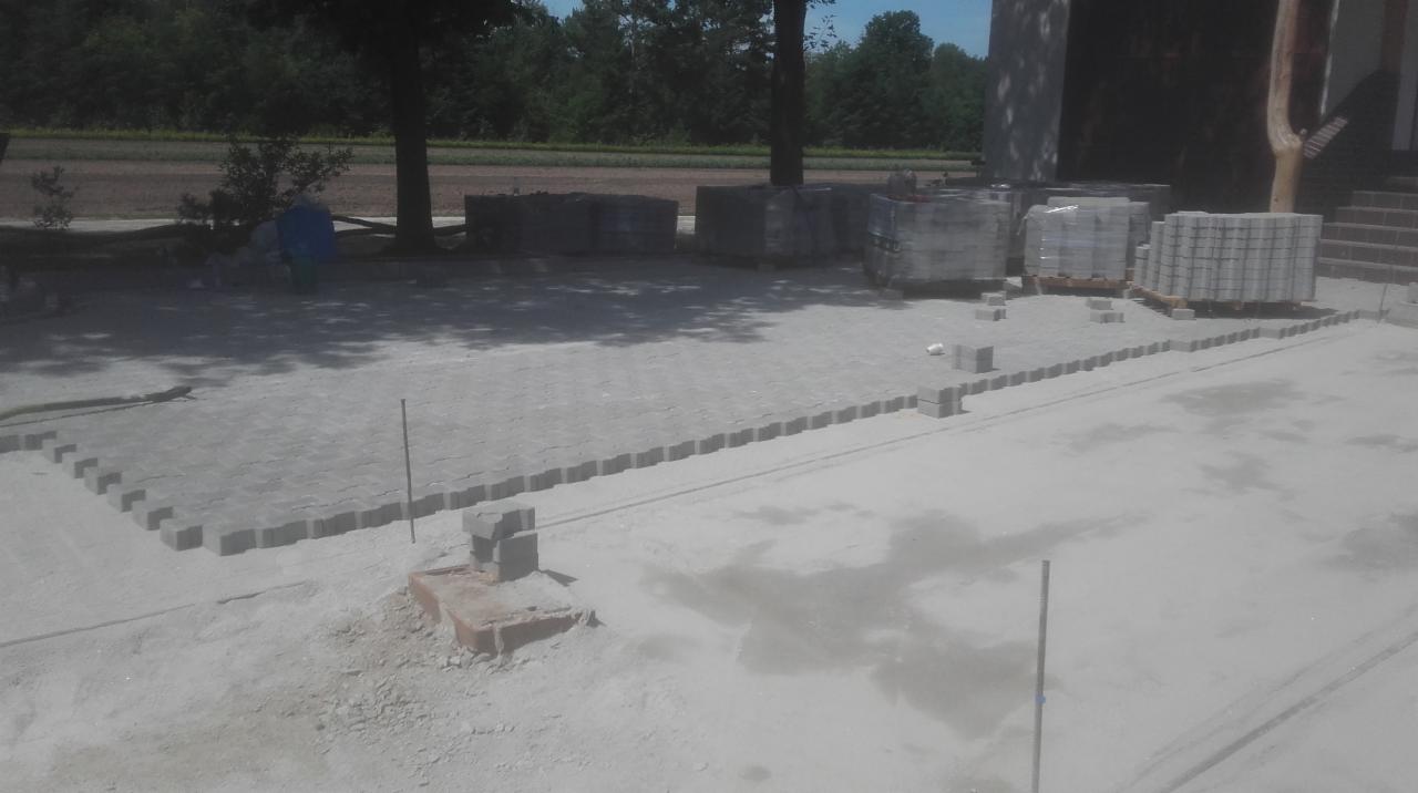 Układanie szarej kostki brukowej na piaskowym podłożu, widoczne palety z kostką w tle, teren budowy.