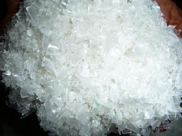 Garść przezroczystych płatków PET w dłoniach, z widocznymi nieregularnymi kształtami i strukturą przypominającą potłuczone szkło.