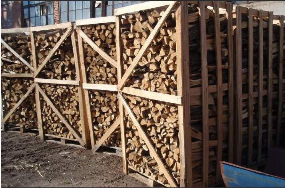 Klatki z surowym, pociętym drewnem kominkowym ułożone na paletach na zewnątrz, z widocznym budynkiem w tle.