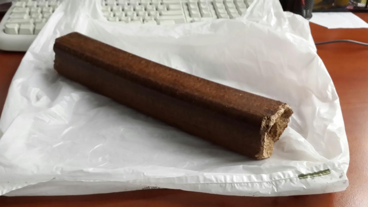 Brykiet drzewny Pini & Kay na białej folii, widoczny ułamany fragment struktury drewna.