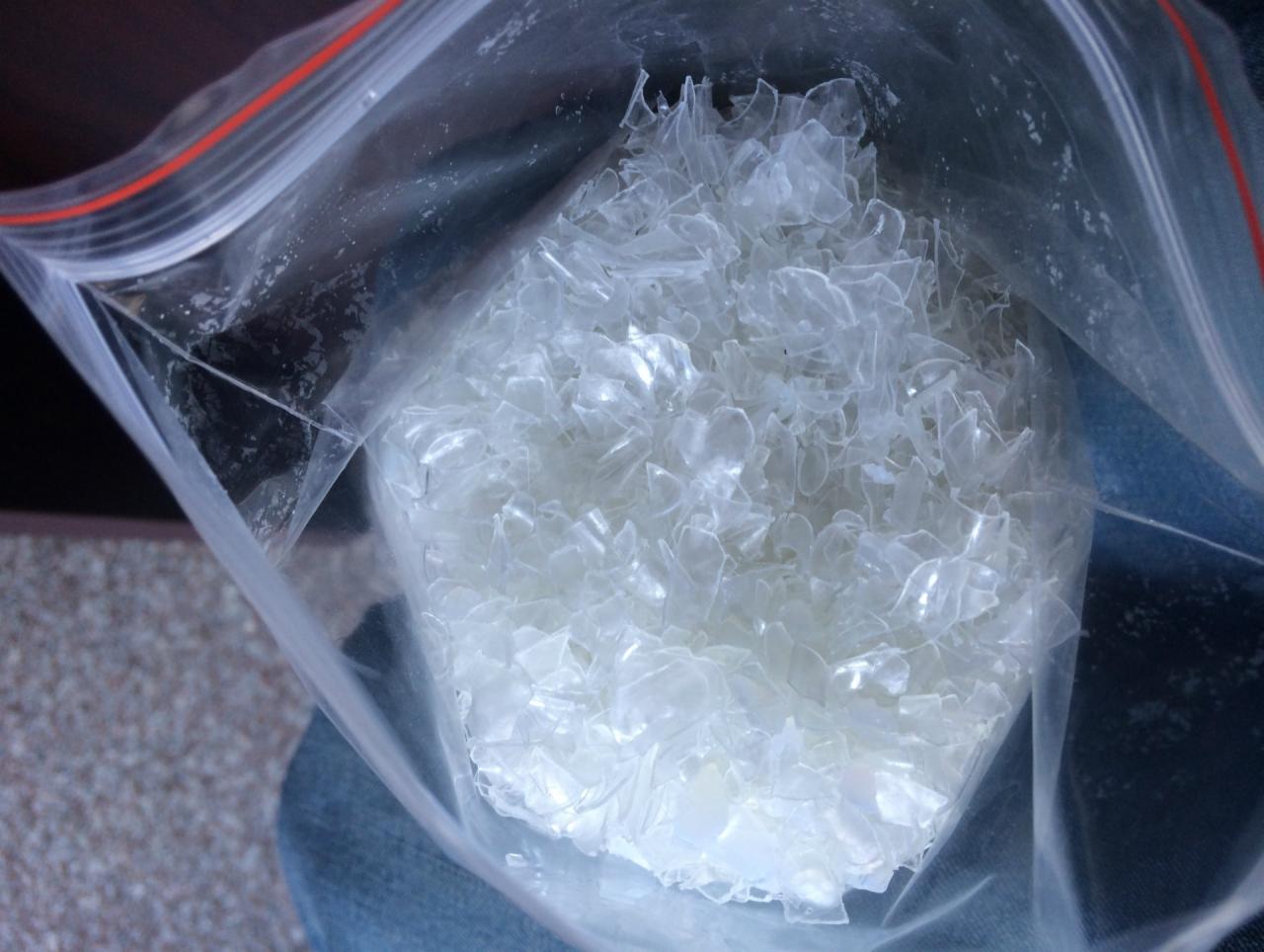 Przezroczysty worek strunowy wypełniony białymi, nieregularnymi płatkami tworzywa PET, widoczne spodnie jeansowe w tle.