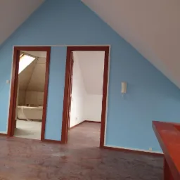 Poddasze po malowaniu: błękitne ściany, drewniane futryny, widok na białą łazienkę i puste, białe pomieszczenie. Podłoga w odcieniach brązu. Skos dachu.