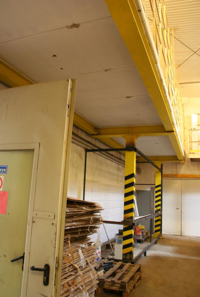 Wnętrze hali przemysłowej z pomostem roboczym o żółtej konstrukcji, widoczne elementy instalacji i składowane materiały budowlane przy ścianie i drzwiach.