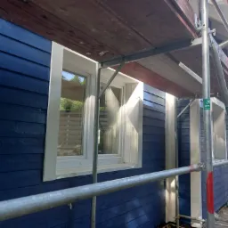 Niebieska drewniana elewacja domu w trakcie prac wykończeniowych, widoczne rusztowanie i białe ramy okienne.