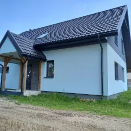 Jasnoniebieski dom jednorodzinny z ciemną dachówką i drewnianym gankiem, widok z zewnątrz na tle błękitnego nieba.