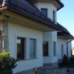 Elegancki dom jednorodzinny z jasną elewacją, brązowymi rynnami i kamienną podmurówką. Widoczne detale architektoniczne i zadbany ogród.