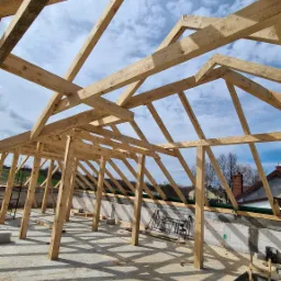 Surowa drewniana konstrukcja dachu w trakcie montażu, widok z dołu na niebo, widoczne elementy więźby dachowej i podłoże z resztkami drewna.