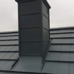 Szary, metalowy komin wentylacyjny z kwadratową nasadą, wkomponowany w pokrycie dachu z blachy trapezowej, na tle pochmurnego nieba.