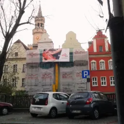 Renowacja kamienicy z rusztowaniami Braas. Widoczne reklamy na siatkach, zaparkowane auta i wieża zegarowa w tle. Czerwona kamienica po prawej stronie.