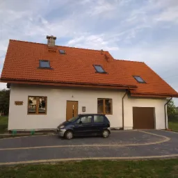 Dom jednorodzinny z czerwoną dachówką, widoczne okna dachowe i komin. Na podjeździe zaparkowany ciemny samochód.