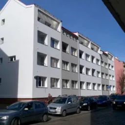 Wielopiętrowy budynek mieszkalny z balkonami, świeżo po odbiorze, z widocznymi detalami architektonicznymi i zaparkowanymi samochodami na ulicy.