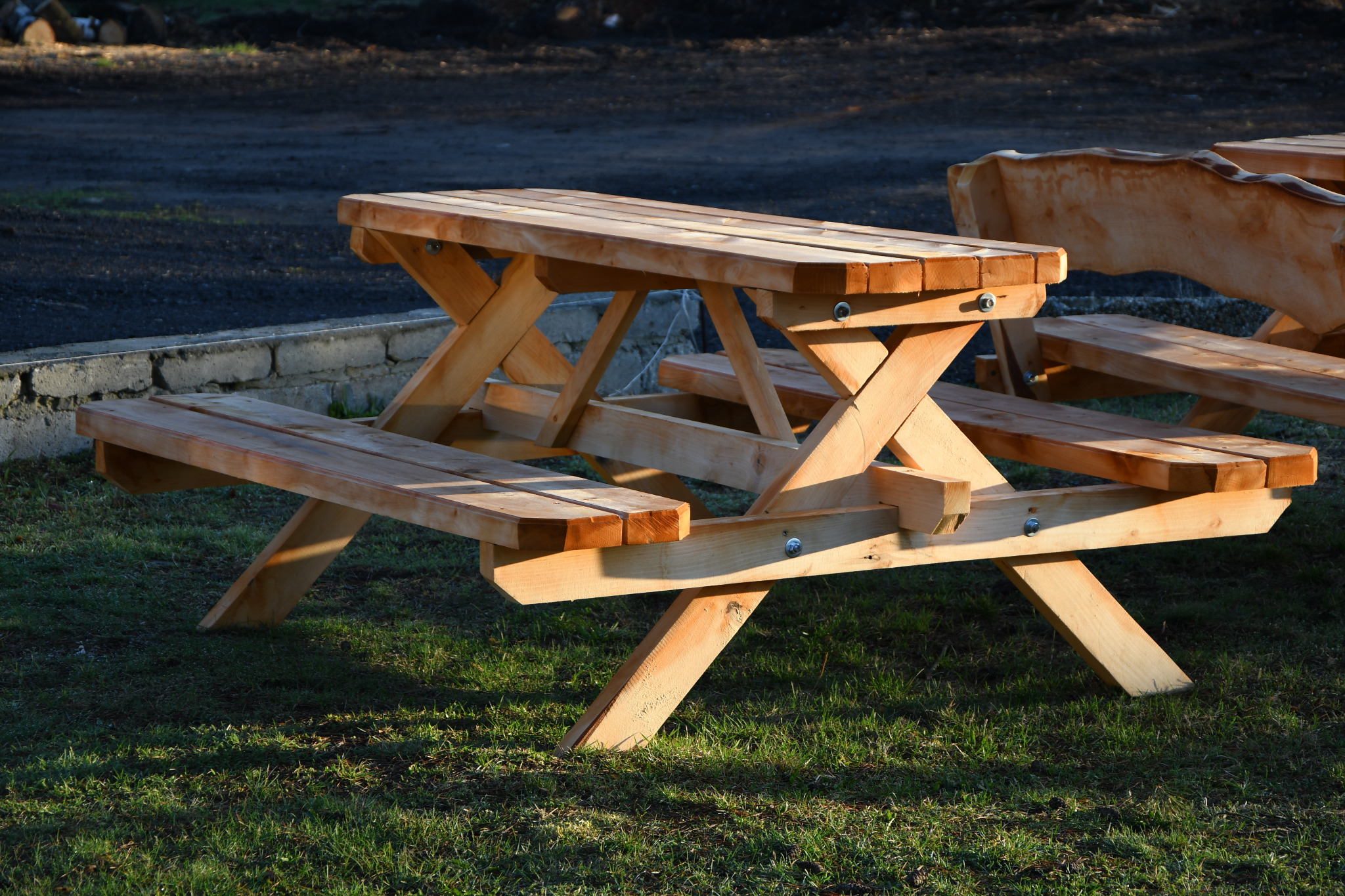 Nowy, drewniany stół piknikowy z ławkami, wykonany z jasnego drewna, ustawiony na trawie w miejscowości Rudy. W tle widoczna droga.