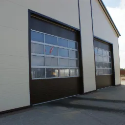 Dwie brązowe bramy garażowe segmentowe z oknami w beżowym budynku przemysłowym, widok z zewnątrz, kostka brukowa, dom w tle, słoneczny dzień.