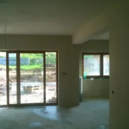 Wnętrze po tynkowaniu: gładkie, jasne ściany z widocznymi oknami i drzwiami balkonowymi. Na zewnątrz widoczny teren budowy i zieleń. Świeżo położone tynki gotowe do dalszych prac.