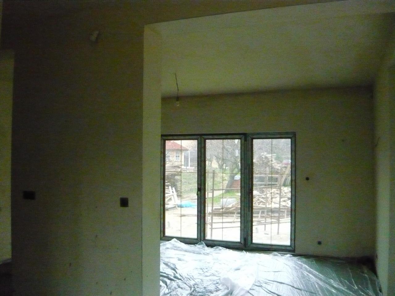 Wnętrze w trakcie prac wykończeniowych: ściany po tynkowaniu, okno trzyskrzydłowe z widokiem na zewnątrz, podłoga zabezpieczona folią malarską.