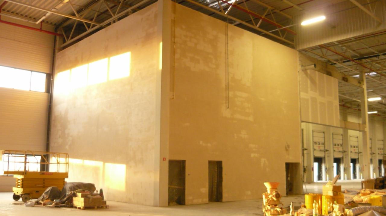 Wnętrze hali z dużą, otynkowaną ścianą działową, widoczne ślady po tynkowaniu, sprzęt budowlany i wpadające światło słoneczne przez okna.