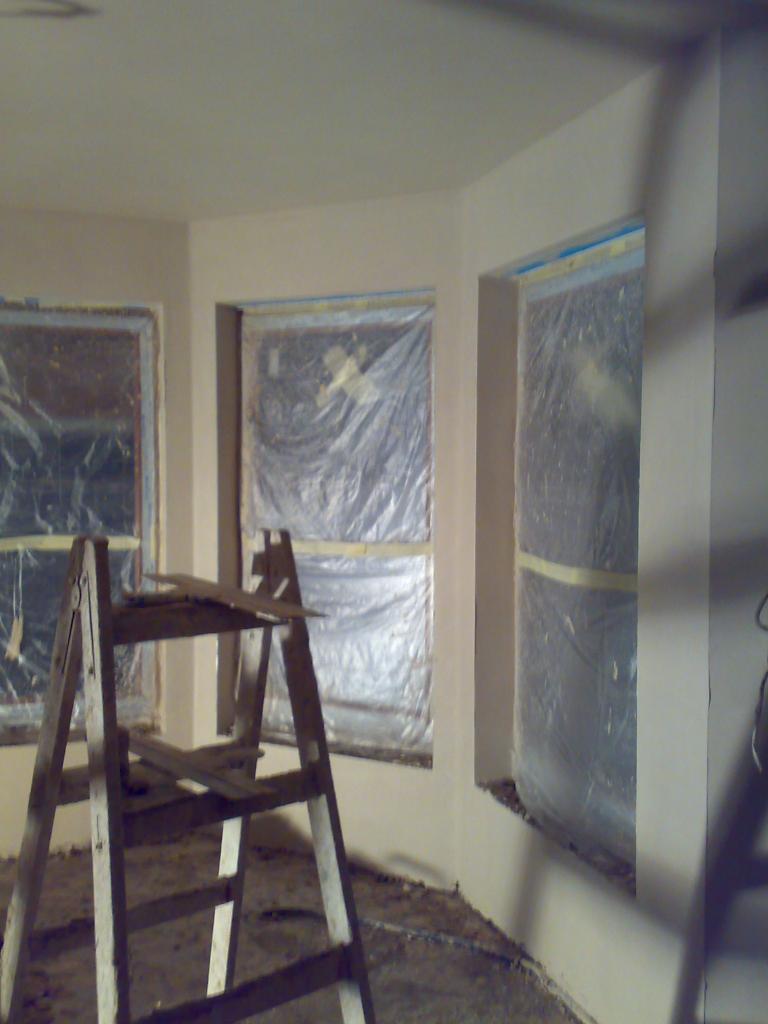 Świeżo otynkowane wnętrze z zabezpieczonymi oknami folią i starą drewnianą drabiną. Widoczne ślady prac na podłodze. Ściany gładkie, gotowe do malowania.