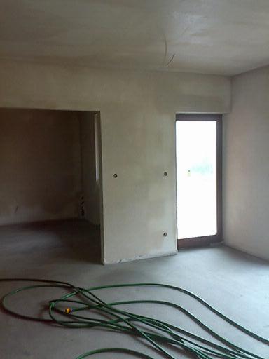 Wnętrze po tynkowaniu maszynowym: gładkie, jasne ściany, widoczne otwory na gniazdka i zielony wąż na podłodze. Surowe wykończenie, przygotowane do dalszych prac.