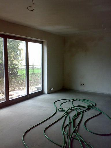 Surowe wnętrze z dużym oknem i zielonym wężem na podłodze. Ściany w stanie surowym, widoczne ślady tynku. Przygotowanie do dalszych prac wykończeniowych.