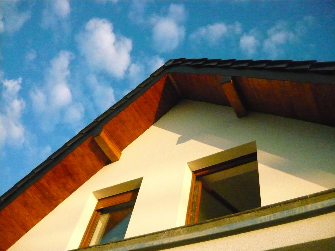 Ściana szczytowa domu z dwoma oknami i drewnianym wykończeniem dachu na tle błękitnego nieba z chmurami. Widok z dołu, ciepłe oświetlenie.