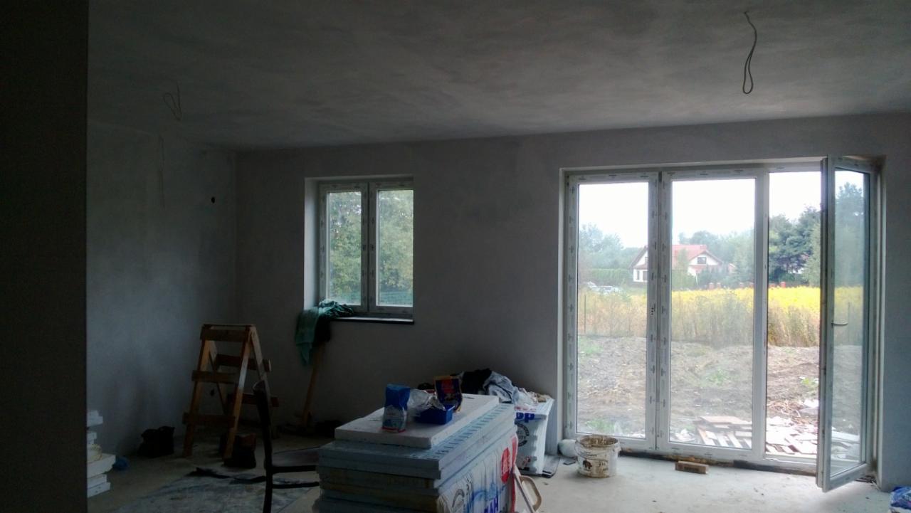 Pokój po tynkowaniu z oknami wychodzącymi na pole. Widoczne ślady prac, drabina i materiały budowlane. Surowy stan wnętrza przed wykończeniem.