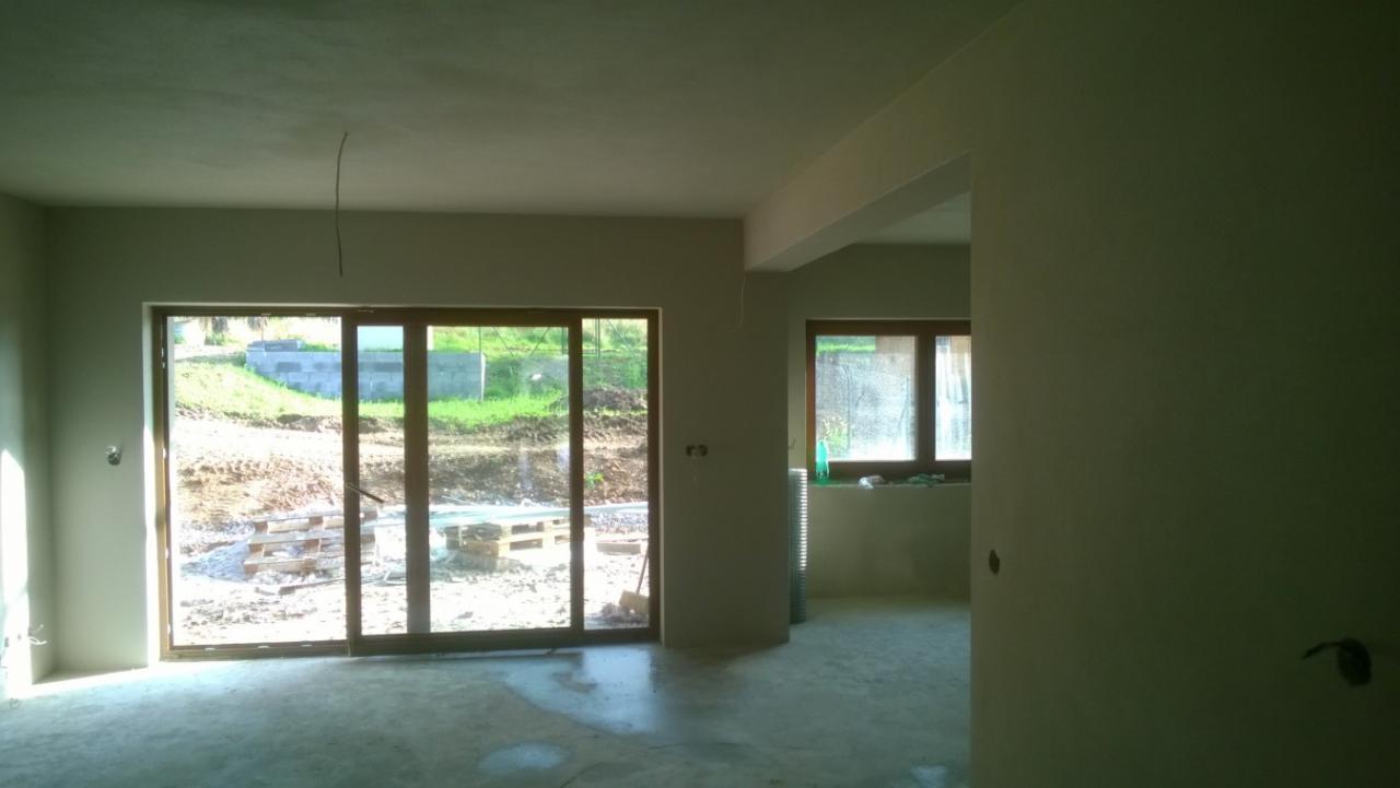 Wnętrze po tynkowaniu: gładkie, jasne ściany z widocznymi oknami i drzwiami balkonowymi. Na zewnątrz widoczny teren budowy i zieleń. Świeżo położone tynki gotowe do dalszych prac.