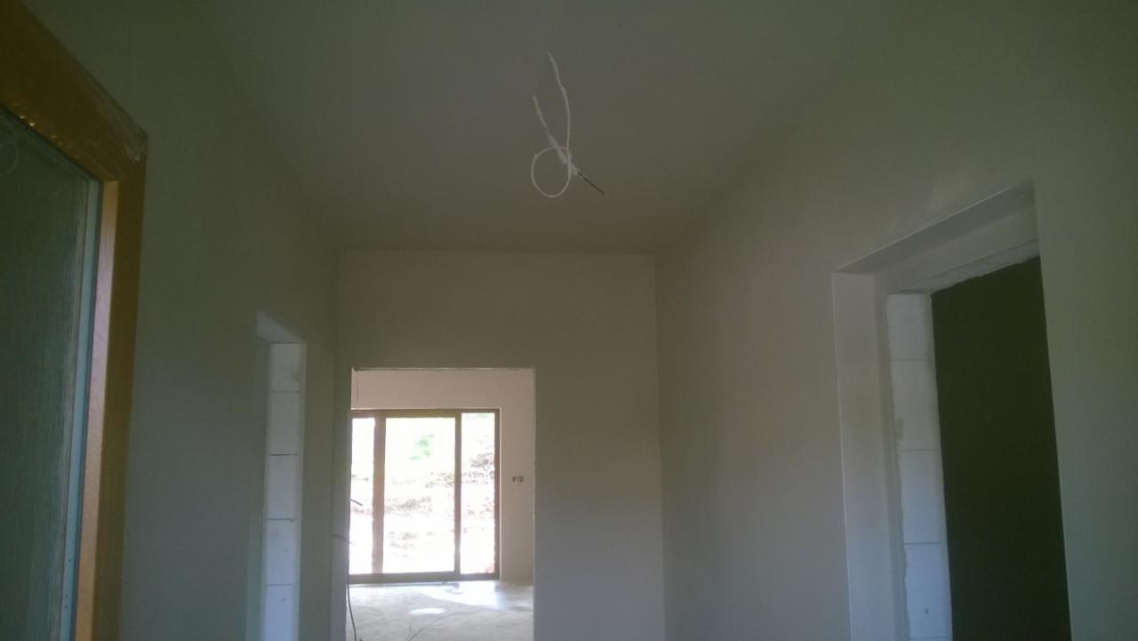 Wnętrze po tynkowaniu: gładkie, jasne ściany i sufit, widoczne otwory drzwiowe i okienne, wystający przewód elektryczny z sufitu, przygotowane do dalszych prac.