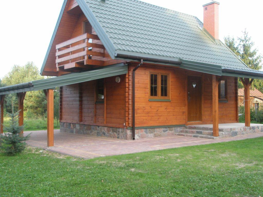 Drewniany dom z bali z zielonym dachem i zadaszonym tarasem w Wiśniew. Widoczny fragment elewacji z oknem i drzwiami wejściowymi, oraz mała choinka.