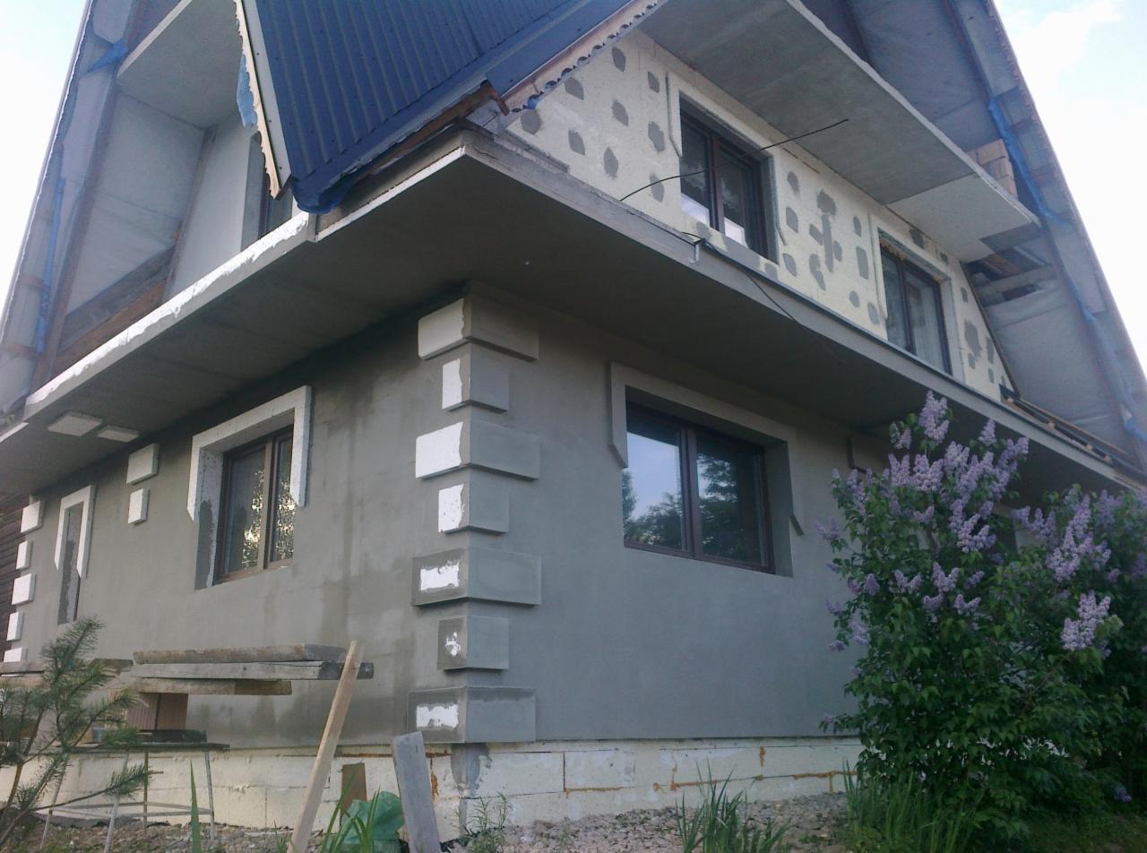 Dom w trakcie tynkowania elewacji z ozdobnymi elementami wokół okien i na narożnikach, widoczne ocieplenie na górnej części ściany, krzew bzu.