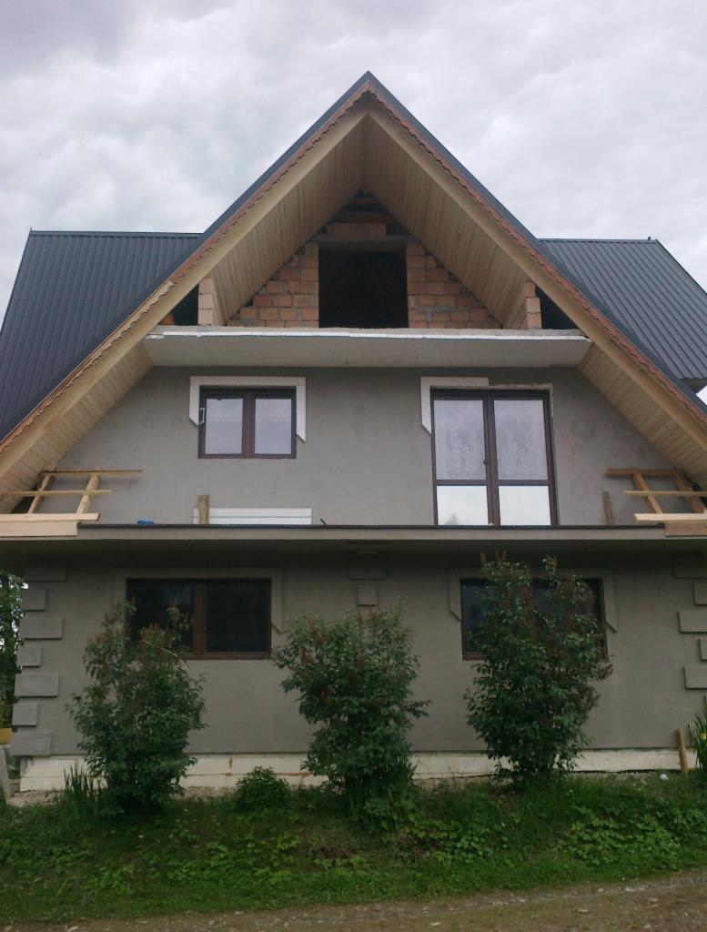 Dom w trakcie budowy z szarą elewacją, widoczne okna, drewniane elementy konstrukcyjne i niezakończona część poddasza z cegły. Prace elewacyjne w toku.