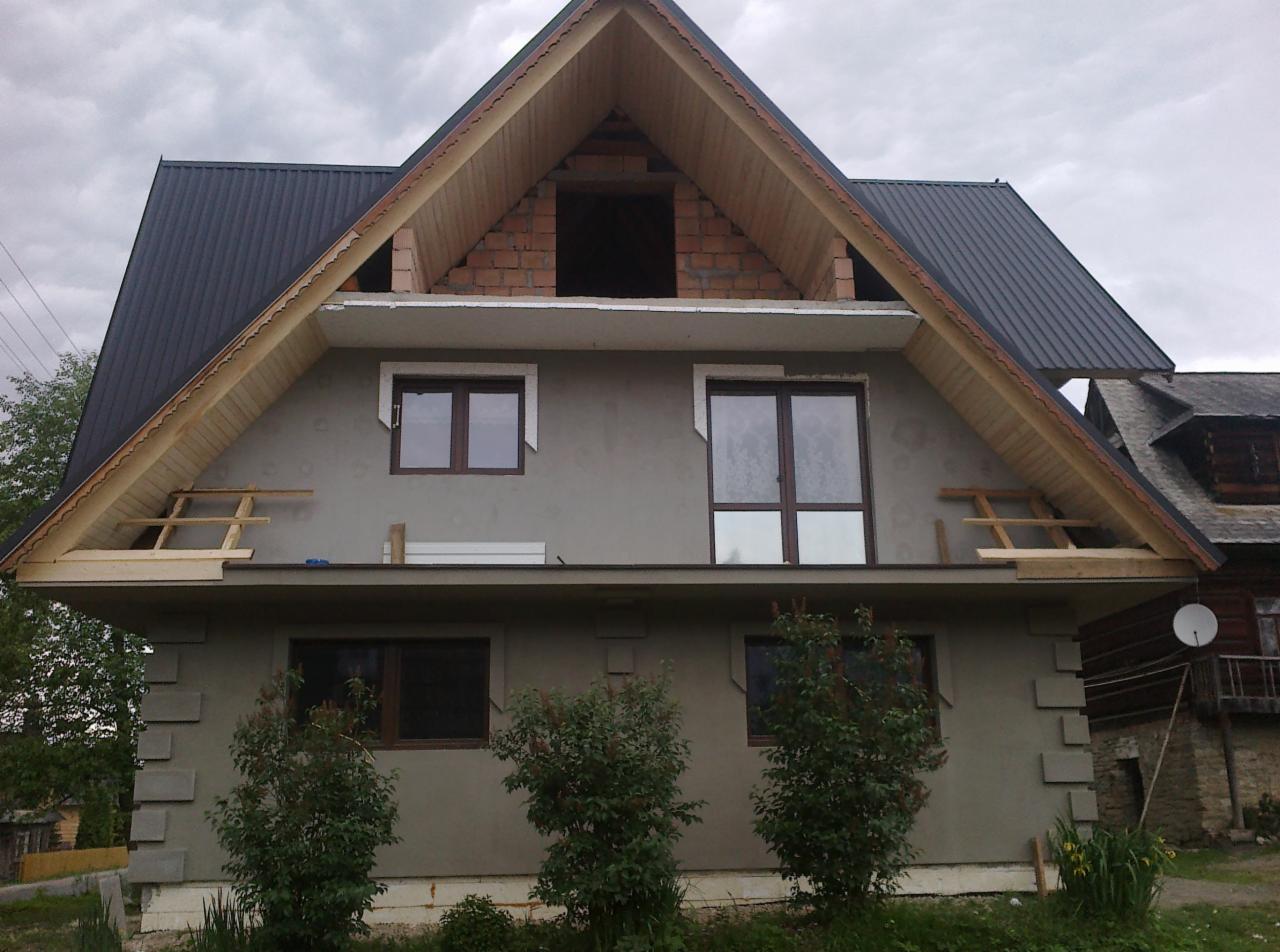 Dom w trakcie ocieplania z widoczną konstrukcją dachu, oknami i surową elewacją. Prace budowlane w toku, widoczne rusztowania i niezakończone detale.