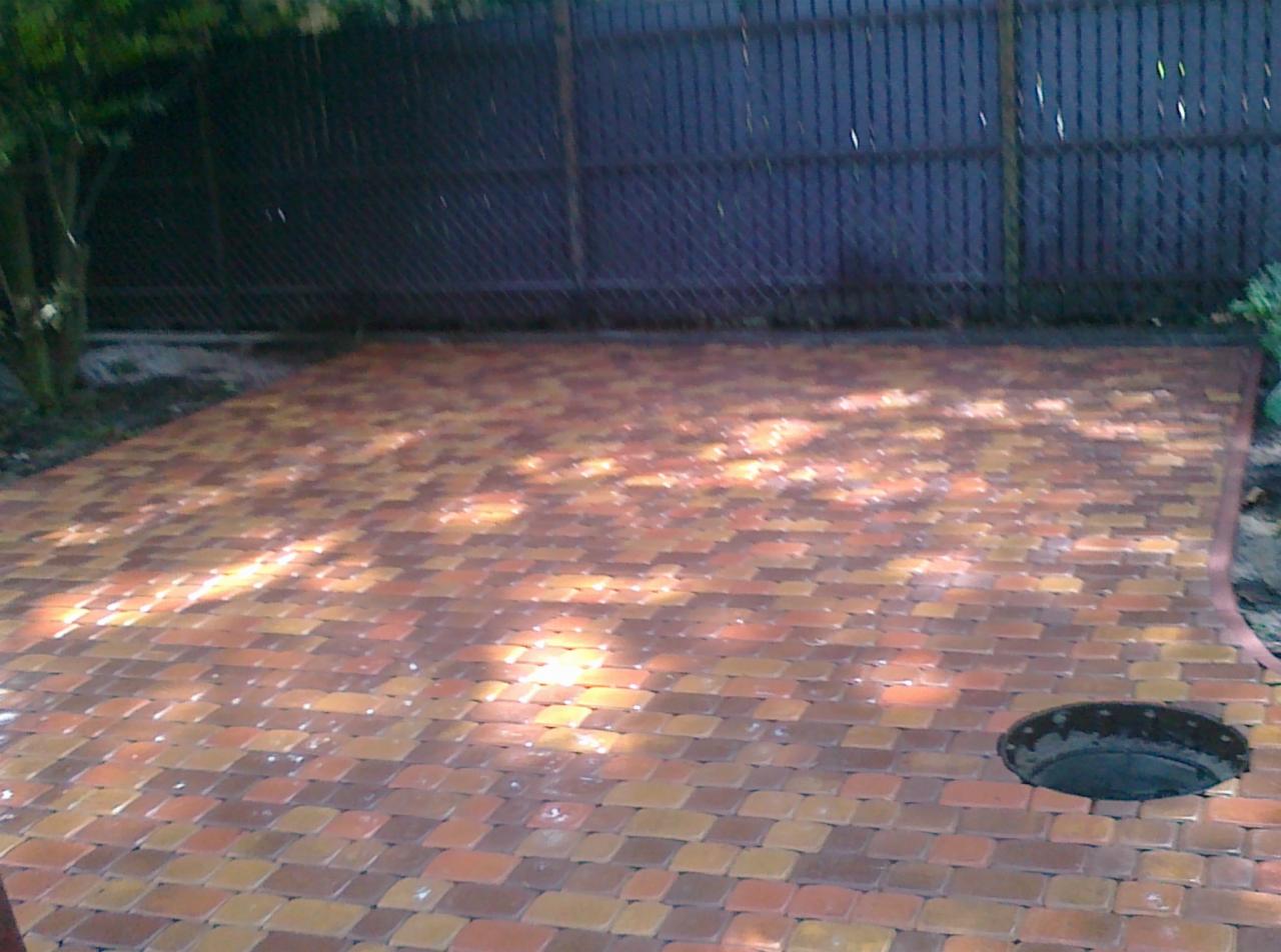 Ułożona nawierzchnia z wielobarwnej kostki brukowej w ogrodzie, widoczny właz kanalizacyjny i fragment ogrodzenia.