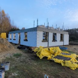 Usługi Ogólnobudowlane Kornosz - Budowa domu jednorodzinnego w stanie surowym otwartym, widoczne ściany z bloczków, zbrojenia na dachu, żółty samochód dostawczy i hałda piasku w tle.