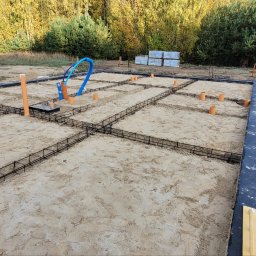 Usługi Ogólnobudowlane Kornosz - Zbrojenie fundamentów pod dom jednorodzinny, widoczne rury instalacyjne, piasek i izolacja pionowa. W tle drzewa i sterta piasku.