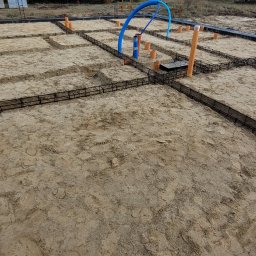 Usługi Ogólnobudowlane Kornosz - Zbrojenie fundamentów pod dom jednorodzinny z widocznymi rurami instalacji, na tle drzew i błękitnego nieba. Widok z góry na przygotowany teren budowy.