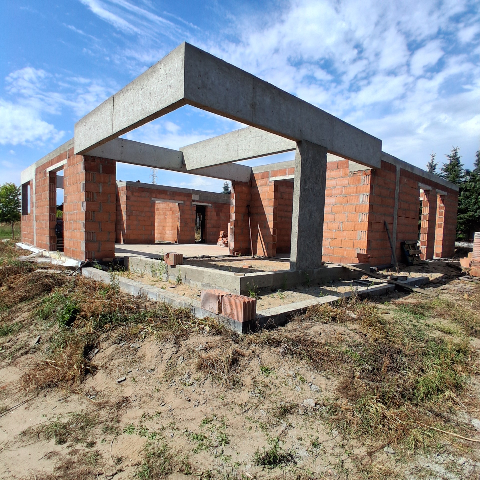 Surowy stan domu z czerwonej cegły z betonowymi nadprożami i słupami, w słoneczny dzień. Widoczne fundamenty i początek ścian. Tło: błękitne niebo.