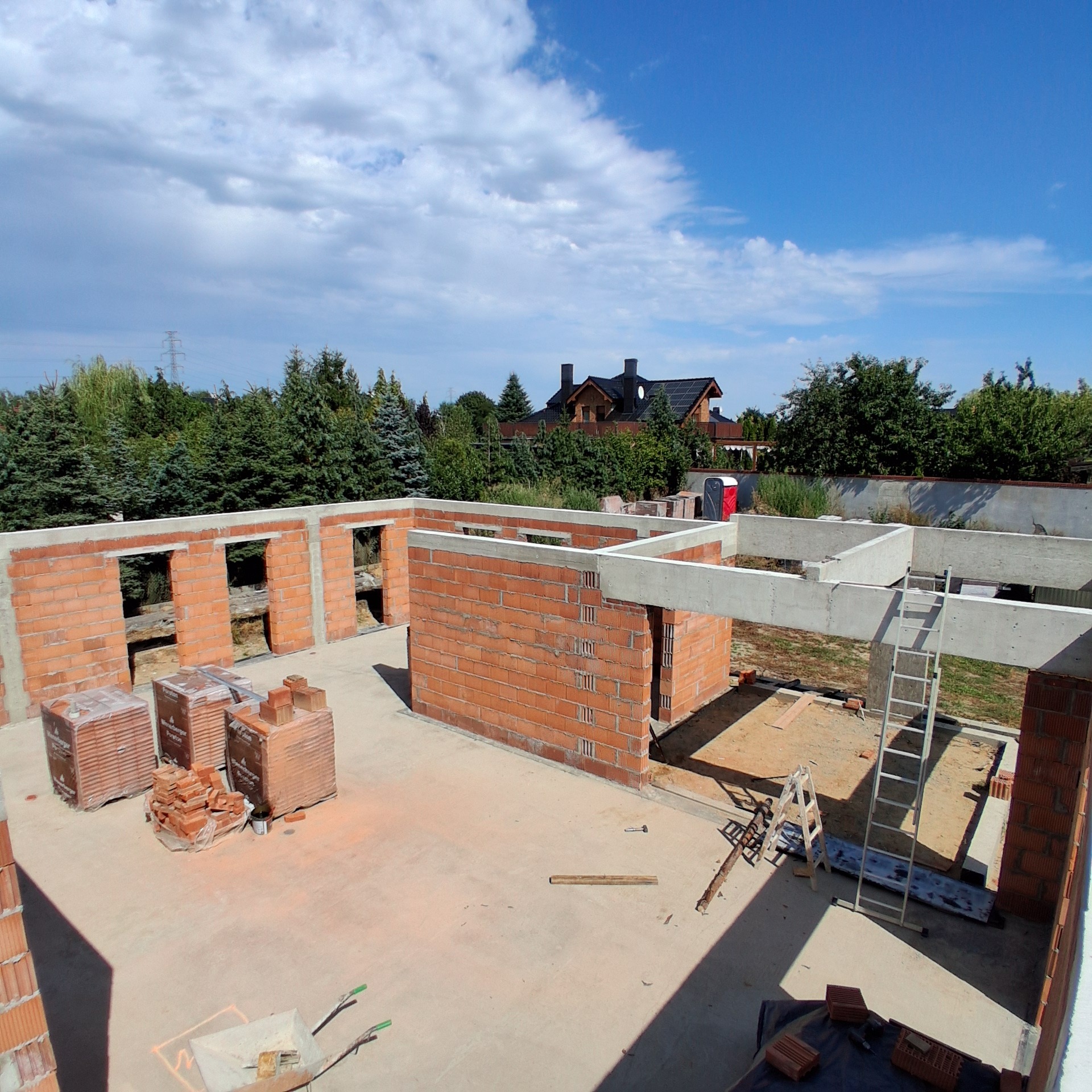 Wysoki kąt na budowę domu z czerwonej cegły Porotherm. Widoczne ściany, betonowe nadproża, palety z cegłami i drabina. W tle dom z ciemnym dachem i zielone drzewa.