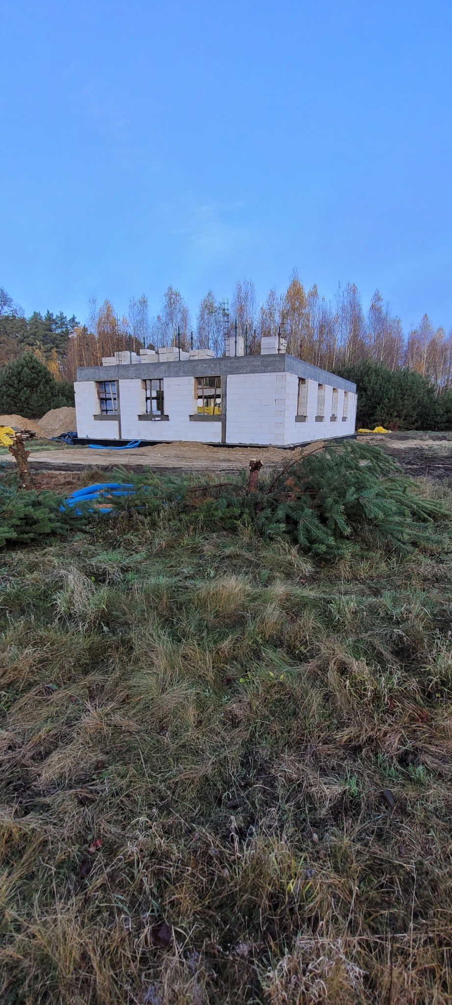 Budowa domu jednorodzinnego w stanie surowym otwartym, ściany z białego betonu komórkowego, widoczne otwory okienne, teren budowy z roślinnością.