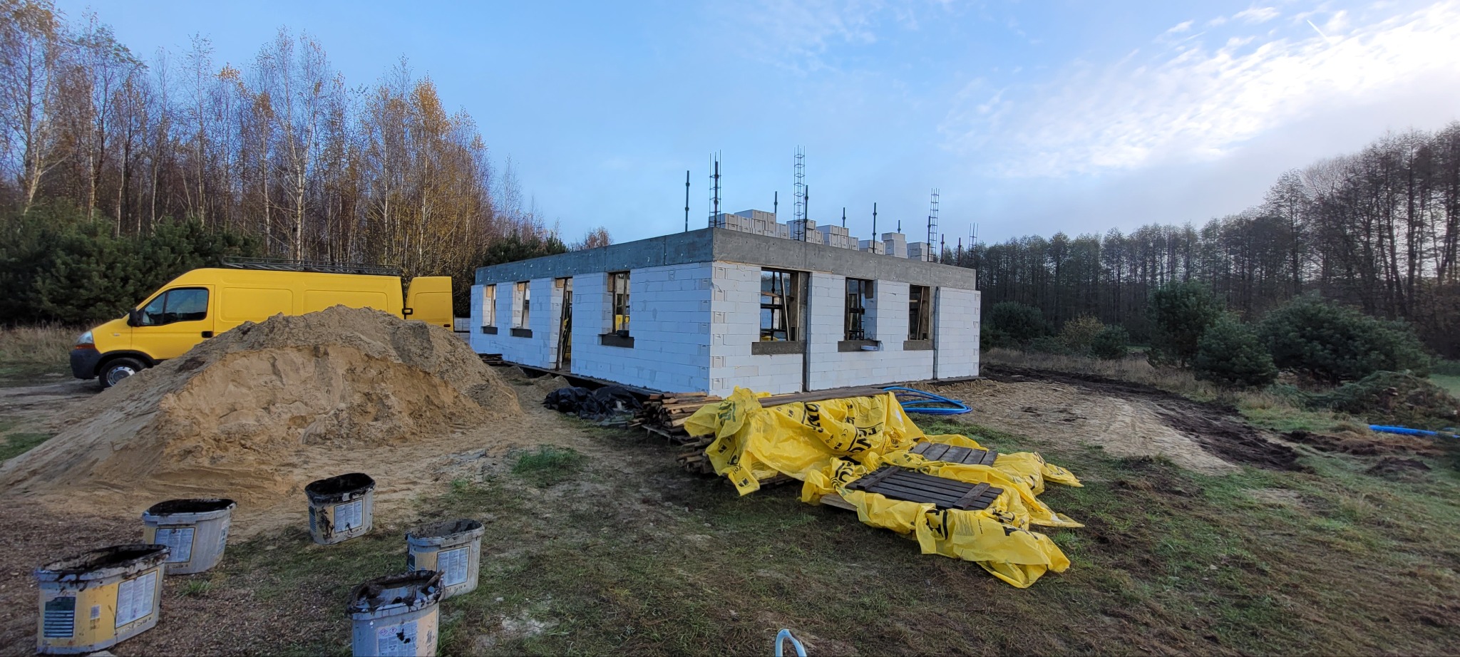 Budowa domu jednorodzinnego w stanie surowym otwartym, widoczne ściany z bloczków, zbrojenia na dachu, żółty samochód dostawczy i hałda piasku w tle.