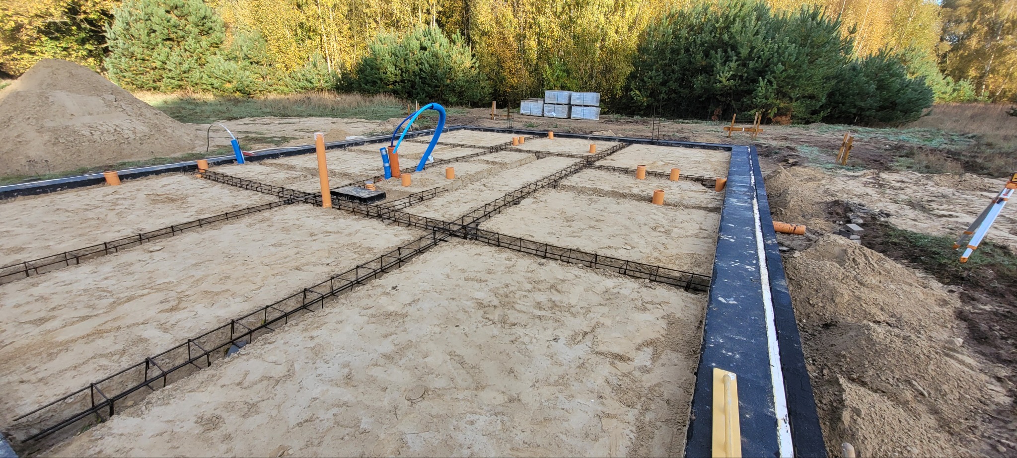 Zbrojenie fundamentów pod dom jednorodzinny, widoczne rury instalacyjne, piasek i izolacja pionowa. W tle drzewa i sterta piasku.