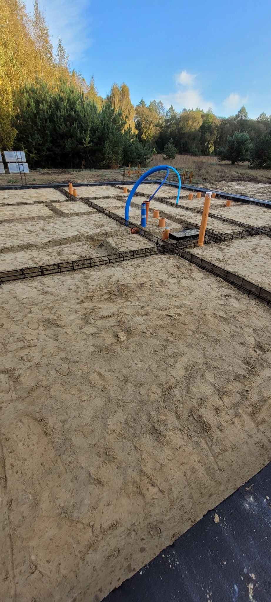 Zbrojenie fundamentów pod dom jednorodzinny z widocznymi rurami instalacji, na tle drzew i błękitnego nieba. Widok z góry na przygotowany teren budowy.