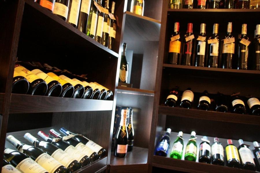 Regały z winami w hurtowni, różne gatunki i roczniki. Butelki ułożone rzędami, etykiety widoczne, oświetlenie podkreśla kolory trunków. Elegancki, ciemny wystrój.